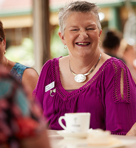Norma sits at a table with a cup of tea smiling and socialising with others.