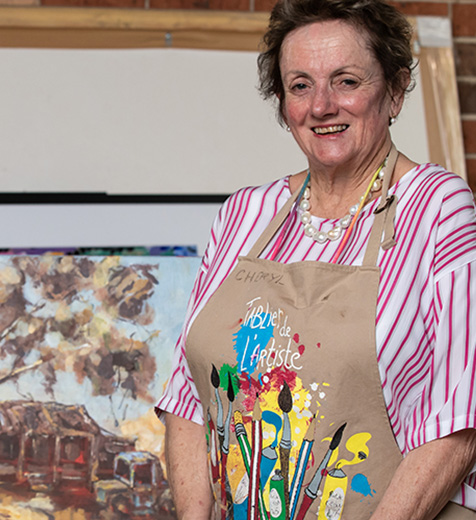 Cheryl dressed in a painters aprons smiles at camera while standing in front of colourful canvases