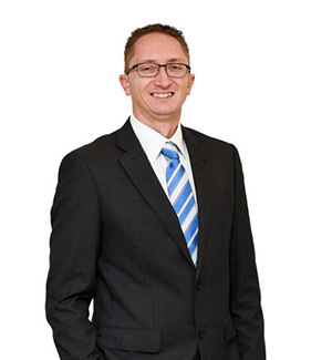 Vision Australia CIO Damien McCormack is smiling at the camera. He has short dark hair and glasses and is wearing a black suit with a blue and white striped tie.