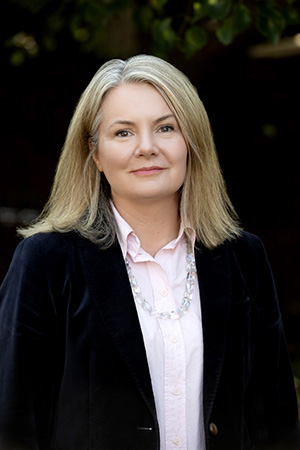 Vision Australia CFO Justine Heath is looking at the camera, smiling slightly. She has shoulder-length blonde hair and is dressed in a dark blazer and pale pink top.